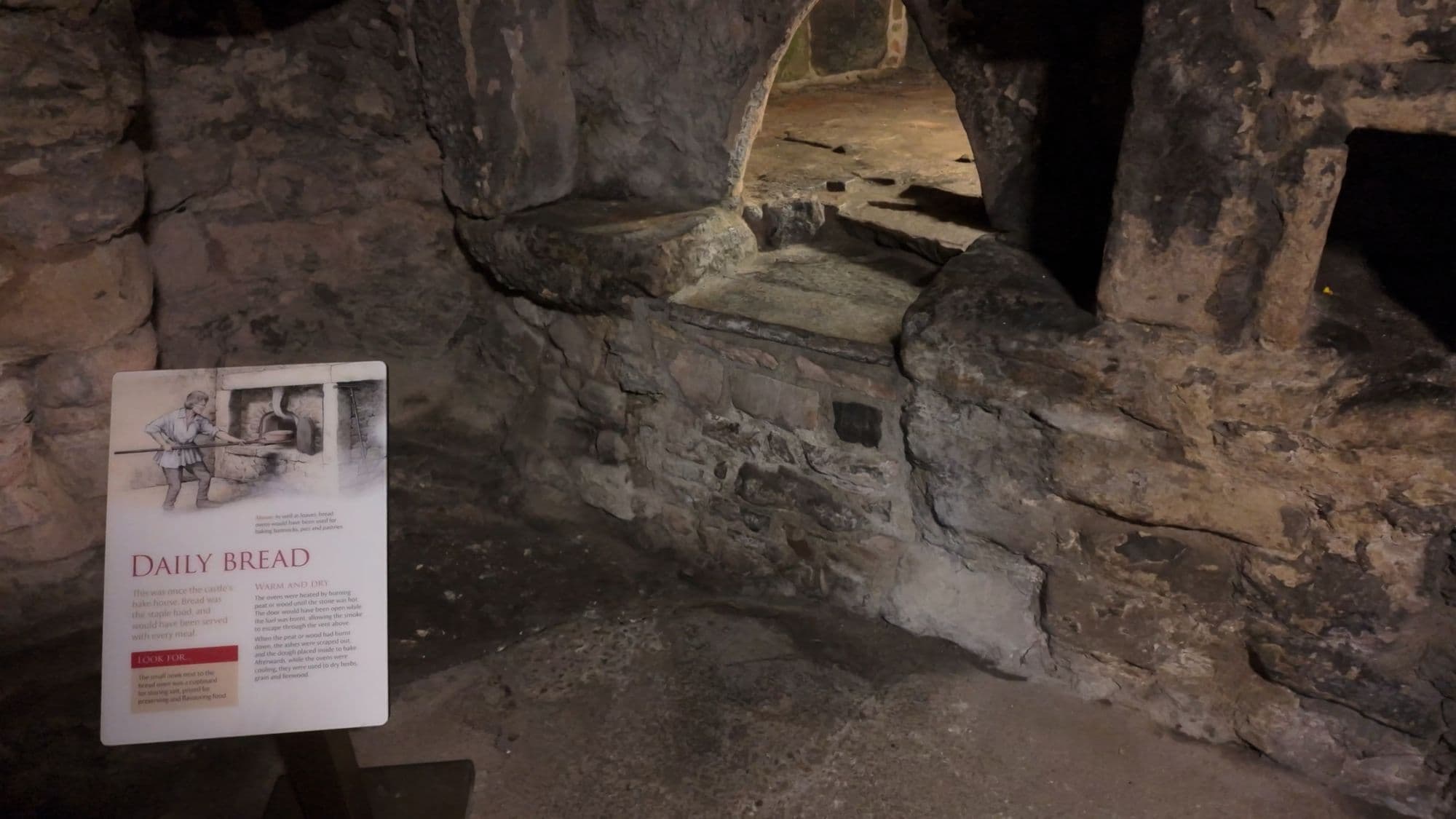 The castle kitchen, with the bread oven still cut into the rock wall beside a 'Daily Bread' interpretation sign