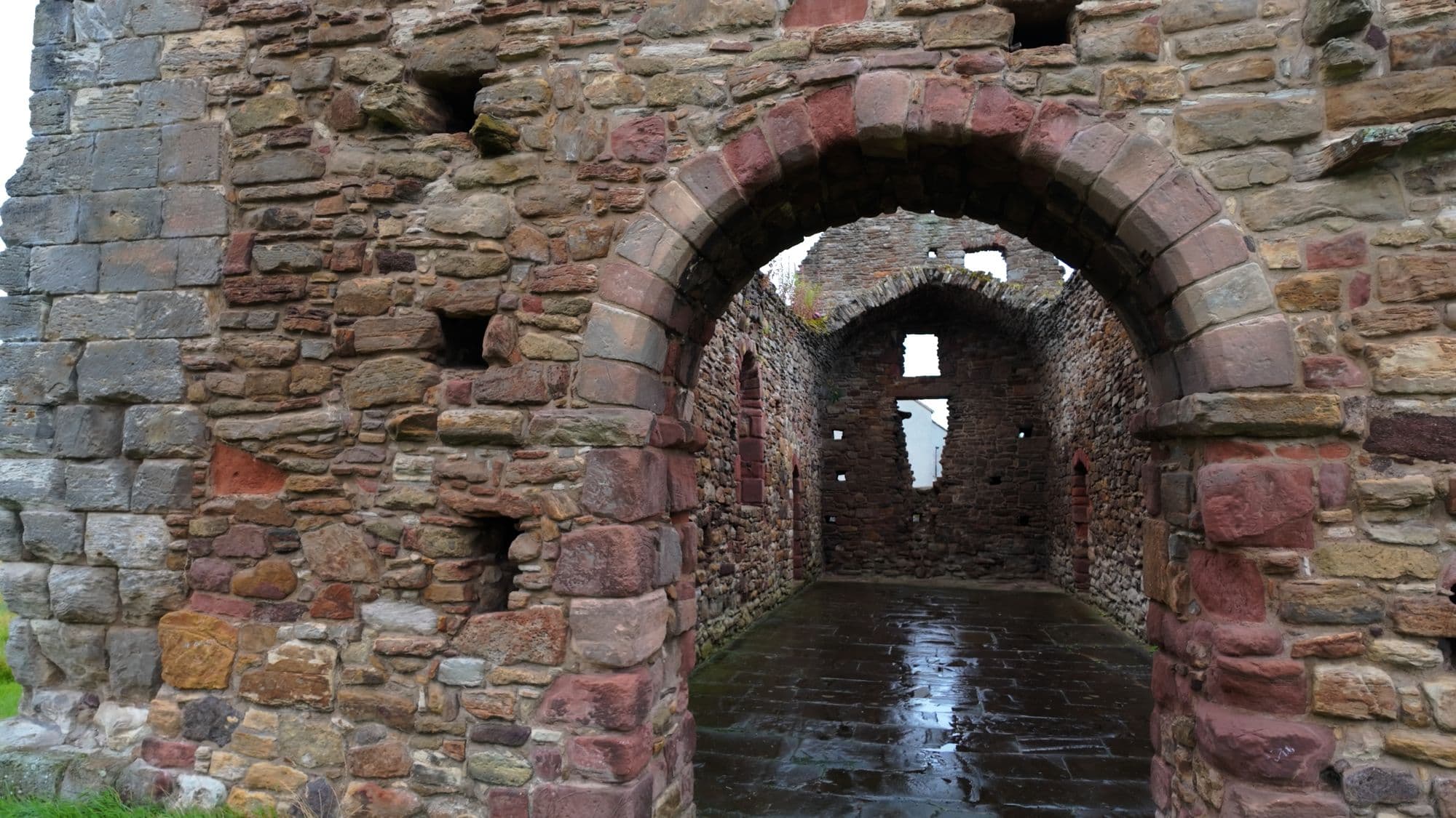 Through a round arch into the roofless nave of St Martin's Kirk, a mix of red and gray stone and a wet stone floor