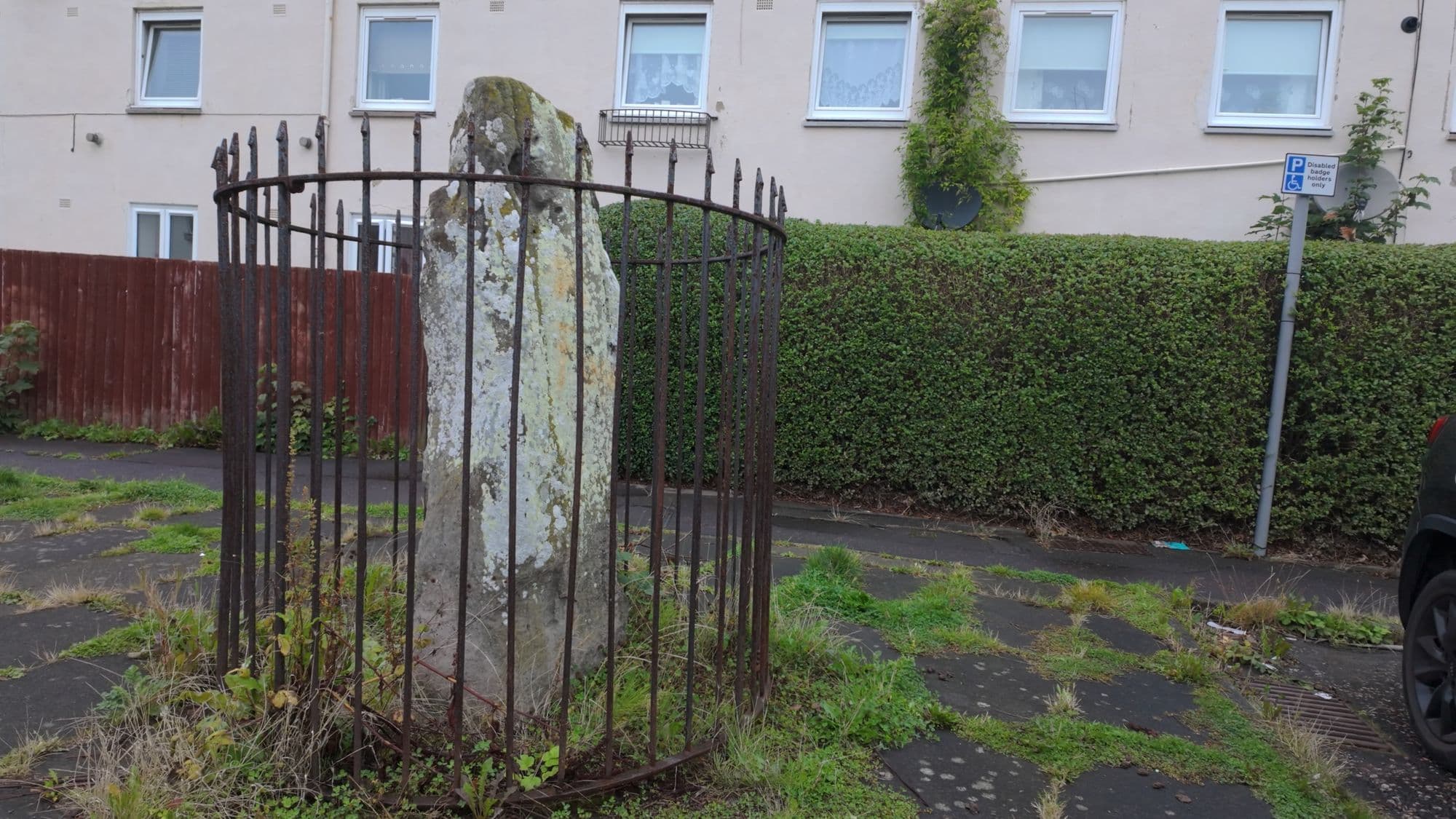 The standing stone in its circular railing on the footpath of the Ravenswood Avenue cul-de-sac, a hedge and housing behind it