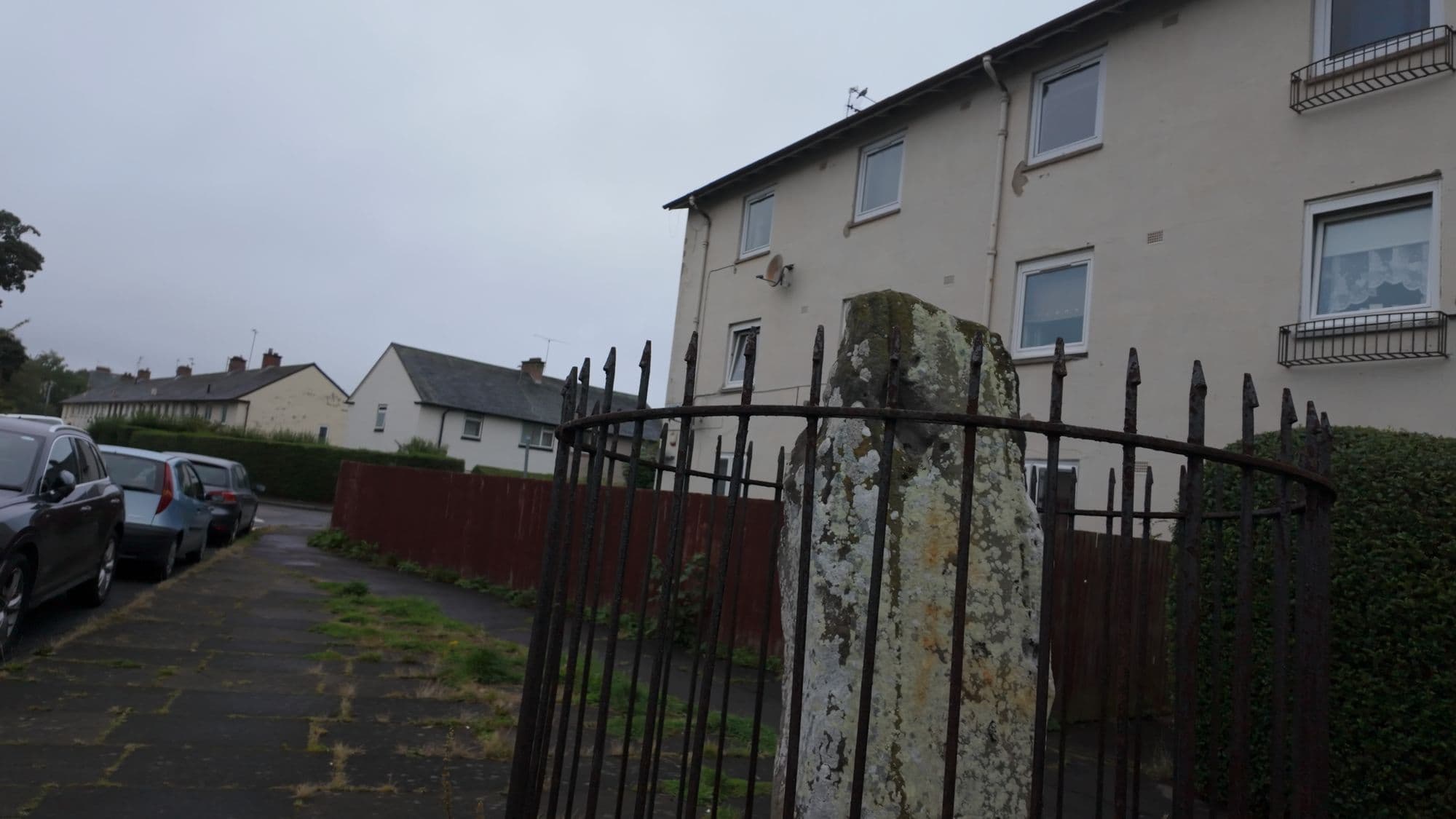 Ravenswood Avenue — a 4,000-year-old stone on the pavement between a fence, a hedge and a row of parked cars