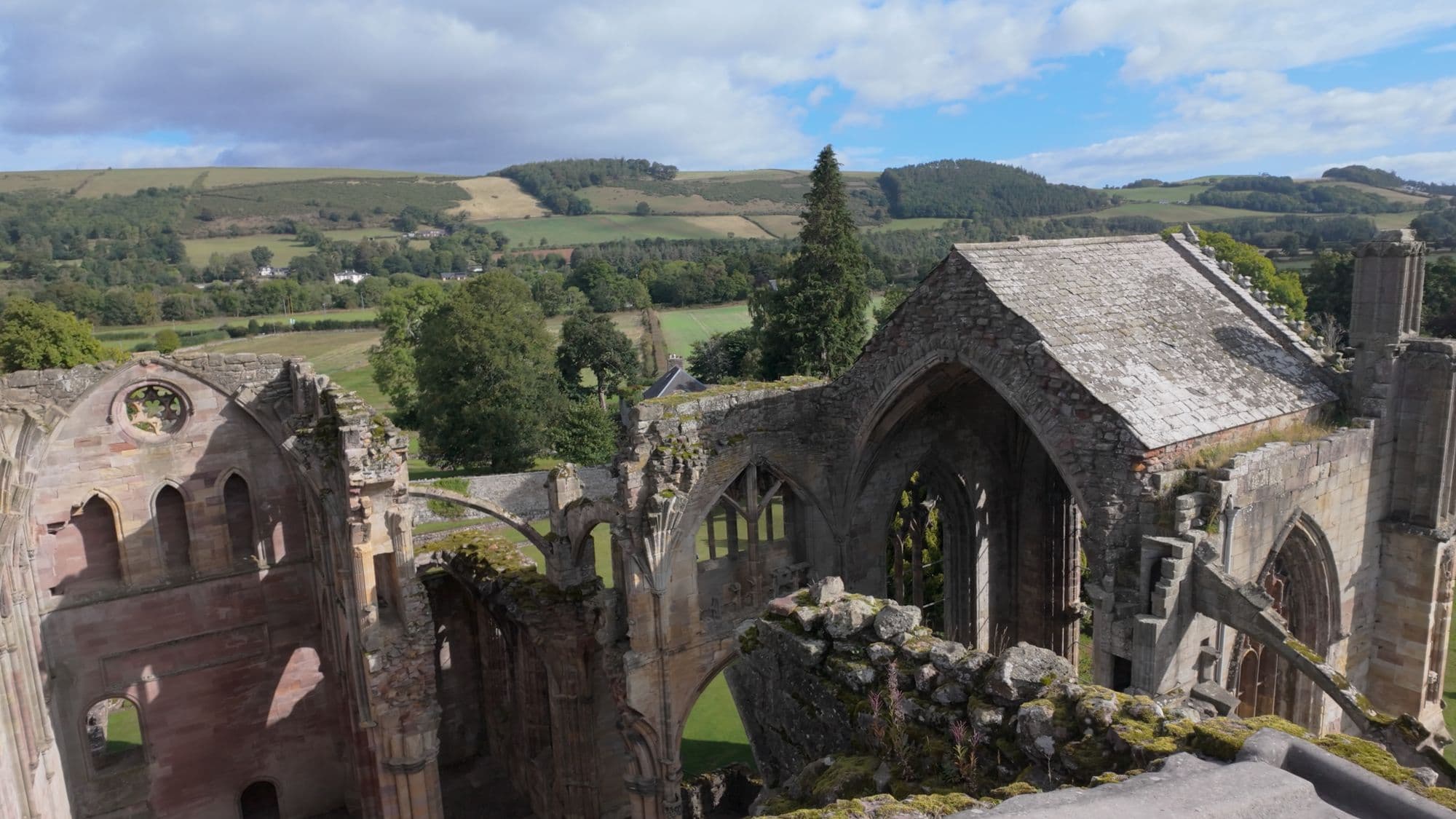 Looking down from high on the abbey walls over the roofless church toward the Tweed valley and the hills beyond