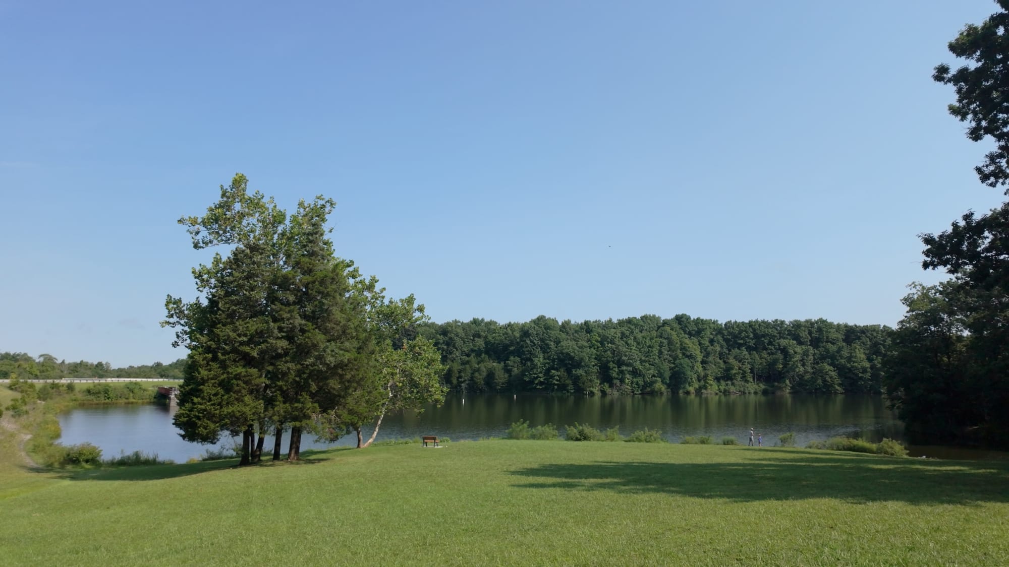 Cowan Lake seen across the mown bank near the dam, with two people fishing from the far shoreline on a clear summer afternoon