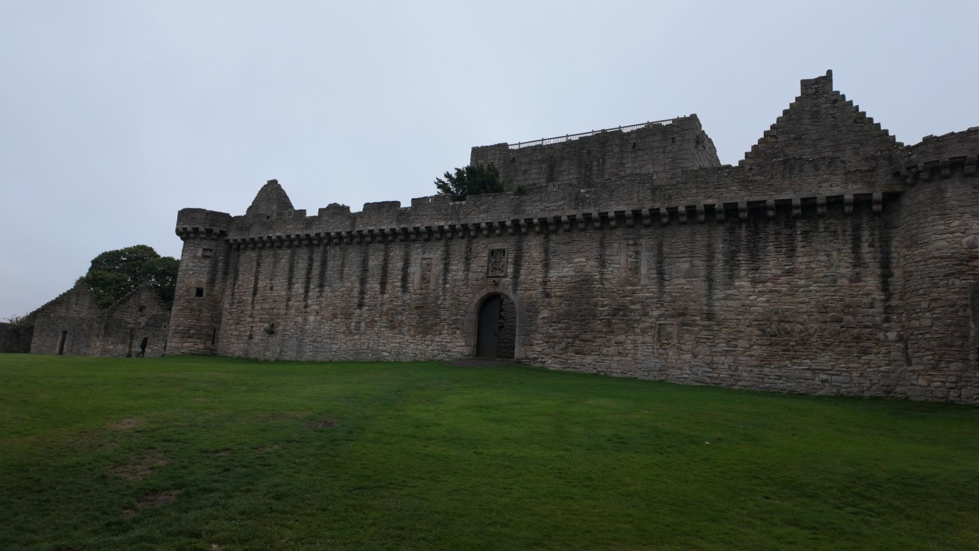 The long battlemented curtain wall of Craigmillar Castle across a wide lawn, the tower house rising behind it under a gray sky