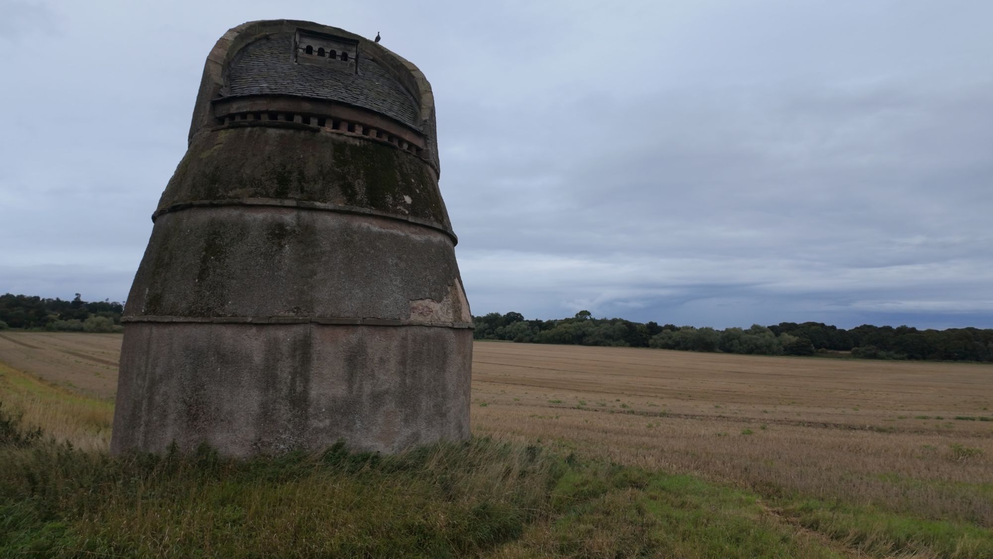 Phantassie Doocot — a squat stone beehive with its top sliced off at an angle, alone at the edge of a harvested field