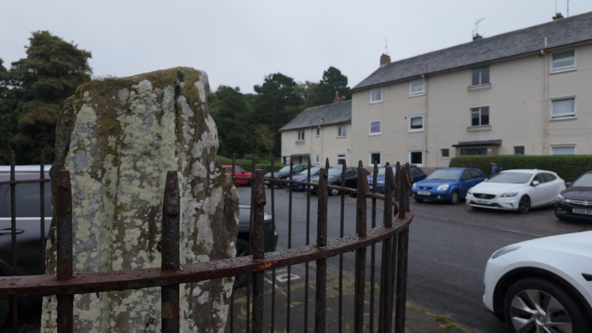 The lichen-covered Liberton standing stone behind its iron railing, with parked cars and a block of flats right behind it