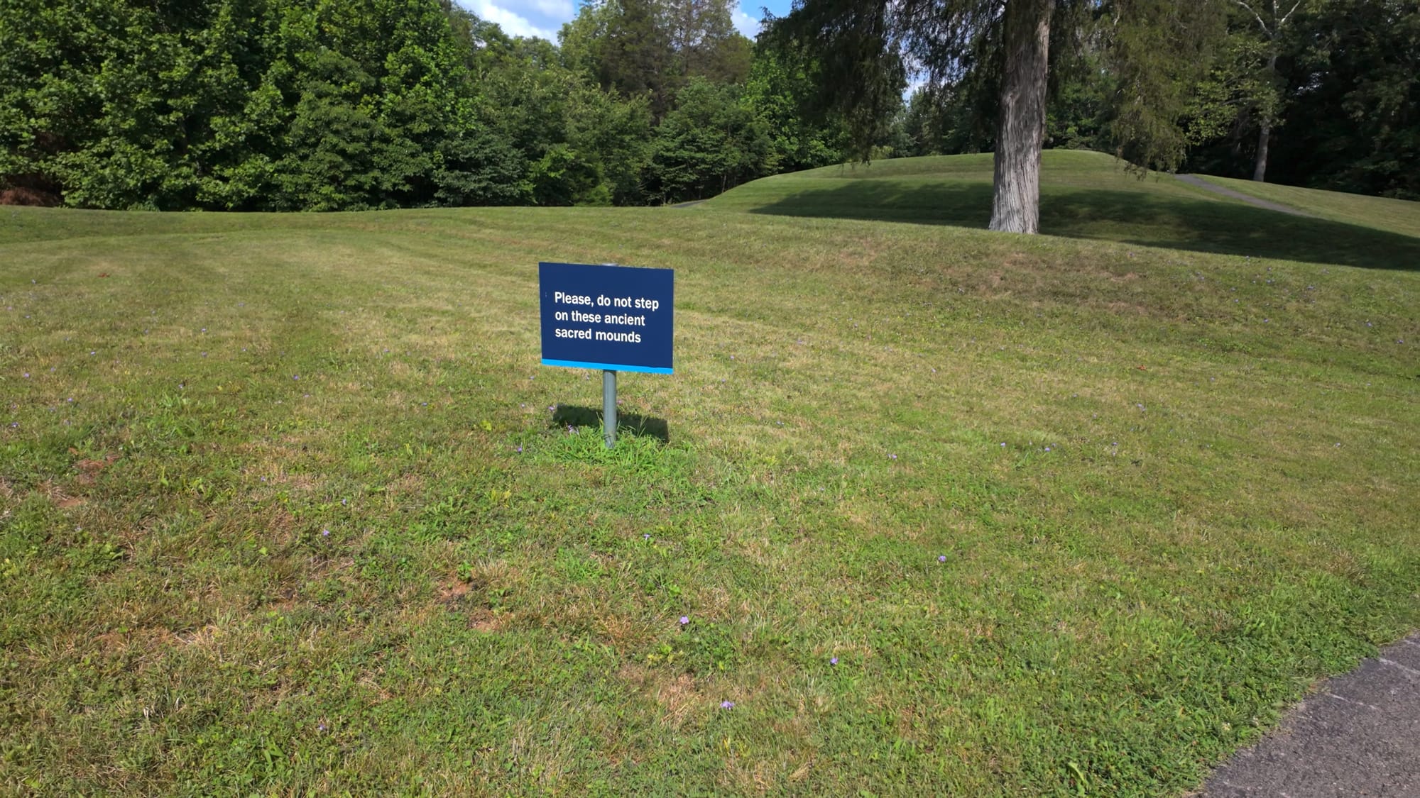 A sign asking visitors not to step on the ancient sacred mounds, with one of the burial mounds visible behind it
